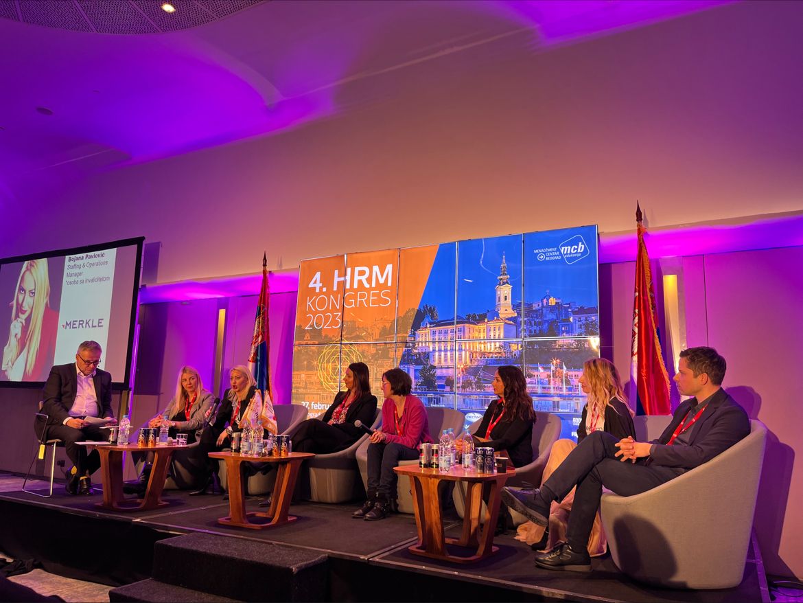 HRM Congress 2023 panel discussion on stage with purple lighting, large cityscape backdrop of Belgrade, and presenter slide of Bojana Pavlović from Merkle. Seven panelists in business attire seated in armchairs.